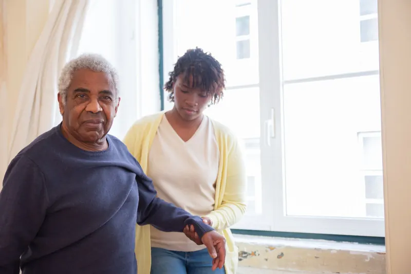 An older man sits by a bright window at home while a caregiver in a yellow cardigan rests a supportive hand on his arm.