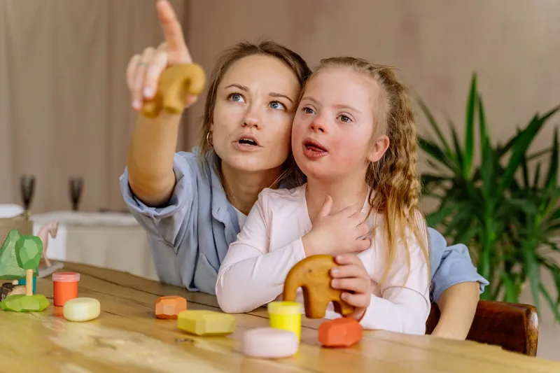 A woman and a girl with Down syndrome play with wooden toys at a kitchen table, the woman pointing at something across the room.