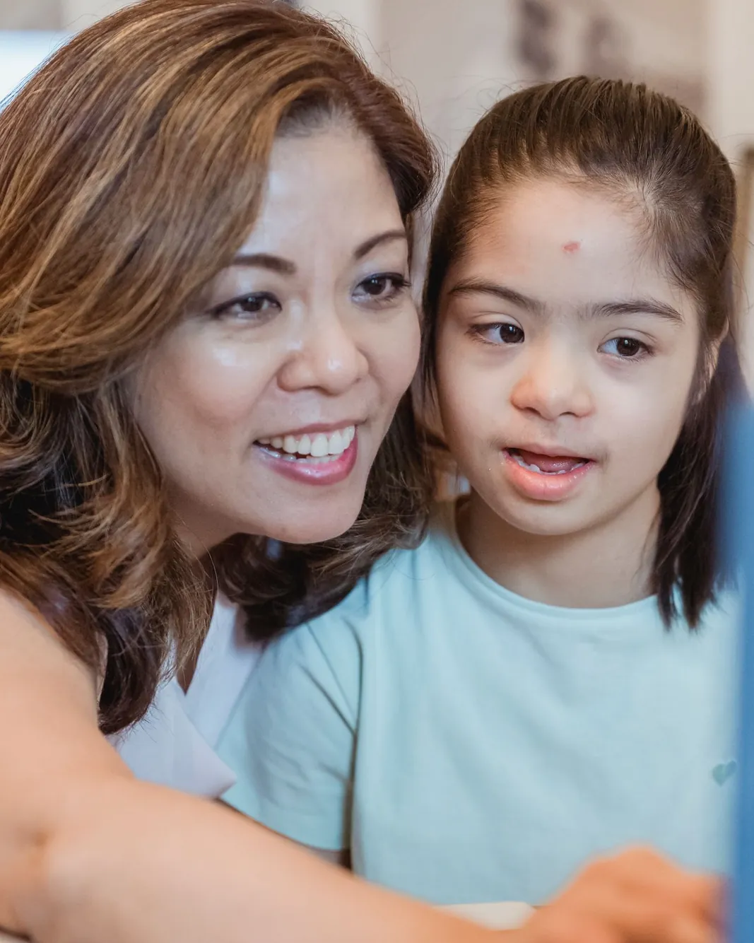 A smiling woman and a young girl with Down syndrome sit close together at a table at home, looking at a laptop.